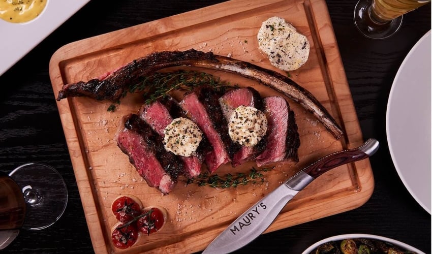 Steak slices with butter on wooden board with knife, herbs, and cherry tomatoes at Maury's